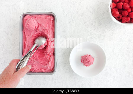 Hand scooping out homemade raspberry ice cream from a metallic tub. Scoop of ice cream in a bowl and fresh raspberries on white marbled background. Stock Photo