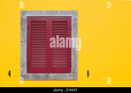 Bright yellow facade with a stone framed grey window, covered with red shutter. Stock Photo