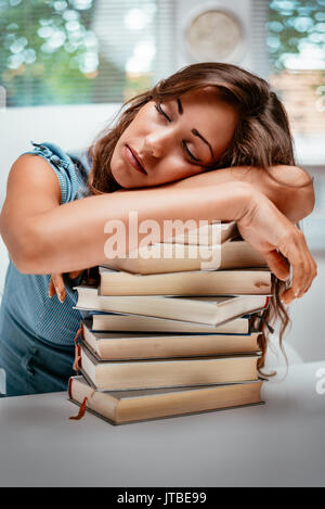 Tired teenage girl sitting in library and sleeping with hands on stack of books. Stock Photo