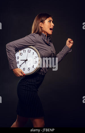 Stressed businesswoman in a hurry running with clock under her arm because she late. Dark background. Stock Photo