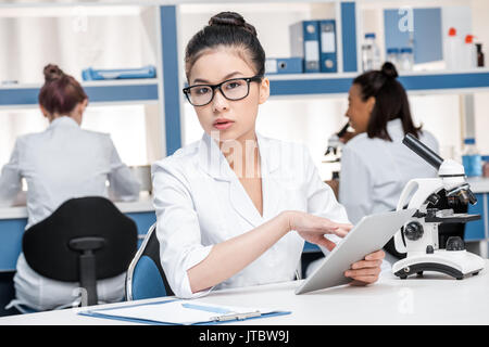 asian scientist in lab coat with microscope and digital tablet working in chemical lab, scientists group behind Stock Photo