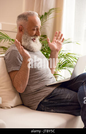 surprised senior bearded man using laptop while sitting on sofa at home Stock Photo