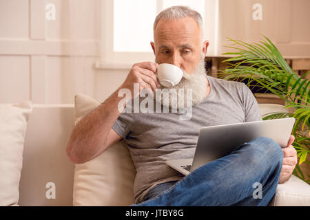 senior bearded man drinking coffee and looking at laptop while sitting on sofa at home  Stock Photo