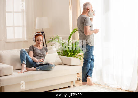 Smiling woman in headphones using laptop and senior bearded man drinking coffee Stock Photo