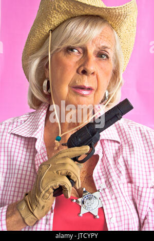 Blond, senior  lady in western outfit: cowboy hat,  deputy sheriff badge on pink checkered shirt, holds black pistol in gloved hand-- against pink Stock Photo