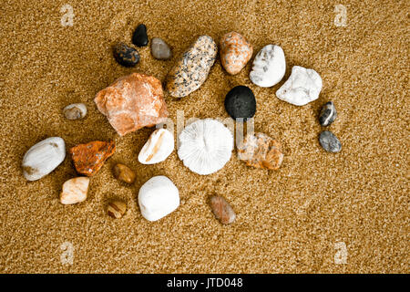 Multi-colored and variegated stones and small rocks gathered from the beach and a sand dollar are shown on a sandy background. Stock Photo