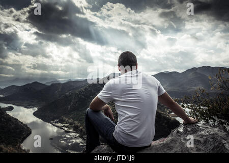 Lonely man looking with hope at horizon on mountain peak with dramatic sunlight during sunset with effect of light at the end of tunnel Stock Photo