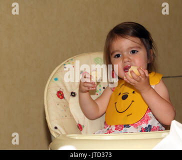 Girl wearing plastic Winnie The Pooh bib while eating bread sitting in high chair England Stock Photo