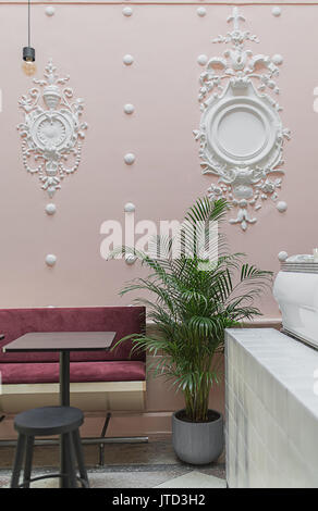 Restaurant with white stucco molding on the pink wall and a  tiled floor. There is a tiled white rack, green plant in the pot, red sofa, black table w Stock Photo