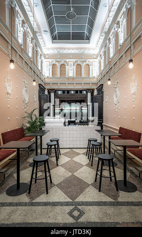 Luminous coffee zone in the antique restaurant with white stucco molding on the walls and large window on a ceiling. There are red sofas, tiled rack w Stock Photo