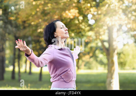 Mature woman runner taking a rest after running in the park. She turn face to sun and hands up. Stock Photo