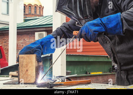 A strong man welder in a construction black overall and welding mask welds a welding machine with a mettalic construction in a wooden beam on the stre Stock Photo