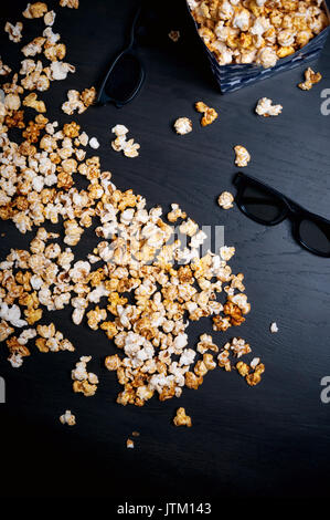 Scattered popcorn on the dark wooden background with couple 3d glasses. Bright movie topic. Fast food. Blue paper box. Watching cinema. Stock Photo