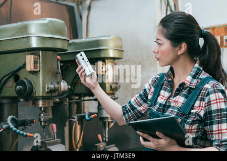 beautiful milling machining employee girl holding mobile digital tablet standing in front of drilling machine and holding components doing inspection. Stock Photo