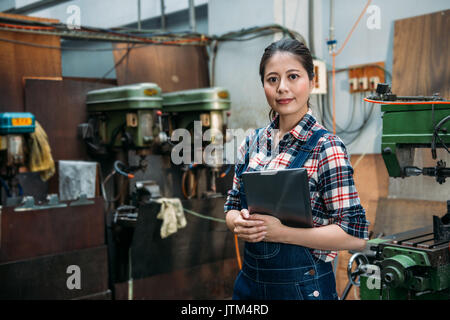 smiling industrial factory woman holding digital tablet computer standing in front of milling machine and confident face to camera. Stock Photo