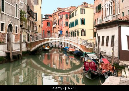 Two Gondolas  in a Venetian canal with a traditional bridge with two people  in the background Stock Photo