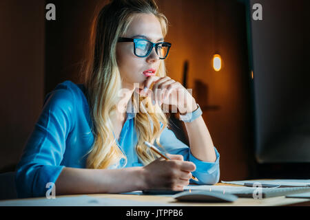 young pensive caucasian businesswoman in eyeglasses writing something with pencil and sitting at workplace Stock Photo