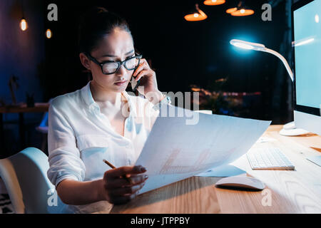 Young asian businesswoman in eyeglasses holding blueprint and talking on smartphone  Stock Photo