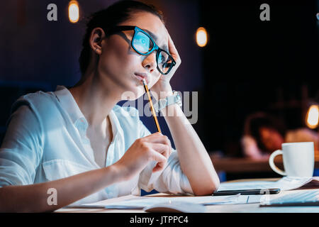 Young businesswoman in eyeglasses holding pencil and working late in office 