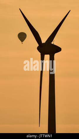 A hot air balloon floating seemingly tightly close to a wind turbine under the colourful light of the setting sun near Lebus, Germany, 09 August 2017. The balloon is actually several kilometers away from the turbine. Photo: Patrick Pleul/dpa-Zentralbild/dpa Stock Photo