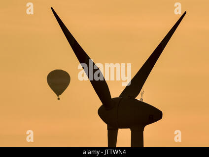 A hot air balloon floating seemingly tightly close to a wind turbine under the colourful light of the setting sun near Lebus, Germany, 09 August 2017. The balloon is actually several kilometers away from the turbine. Photo: Patrick Pleul/dpa-Zentralbild/dpa Stock Photo