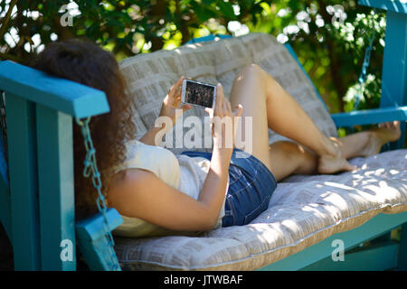 Young woman lying on a patio bench with an iPhone in her hands, watching a movie on the phone screen Stock Photo