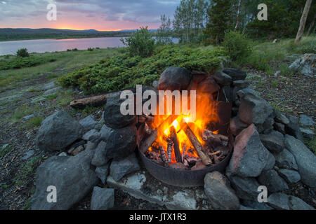 A campfire burns hot at a lakeshore campsite, as the sun sets in the mountains of Wyoming. Stock Photo