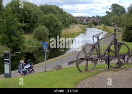 mother and child both pushing prams  pass the Bankies bike sculpture at Clydebank    in Summer weather on the Forth and Clyde canal towpath NCN 7 Stock Photo