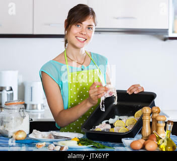 Smiling woman preparing merluccid hake on baking sheet at home Stock Photo