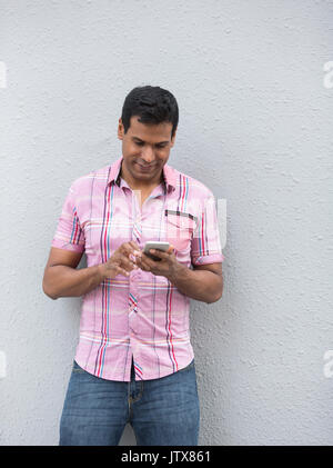 Happy Indian man using a smart phone and leaning against a grey wall. Stock Photo