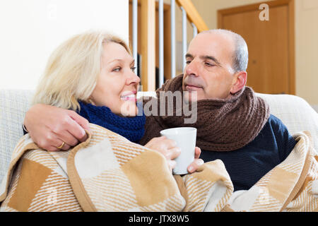 Mature positive spouses under blanket drinking tea on couch Stock Photo