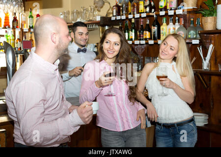 Handsome man talking to two beautiful caucasian women in a bar while smiling bartender is mixing cocktails in the background Stock Photo