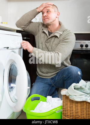 Man is holding his head sitting near washing machine thinking of how to use it. Guy doing laundry Stock Photo