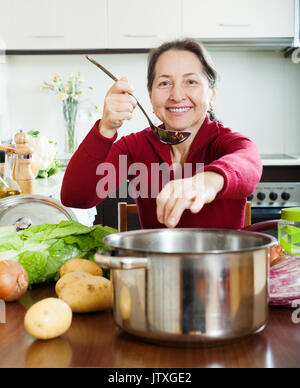 Smiling mature woman adding salt in to soup pan in home kitchen Stock Photo