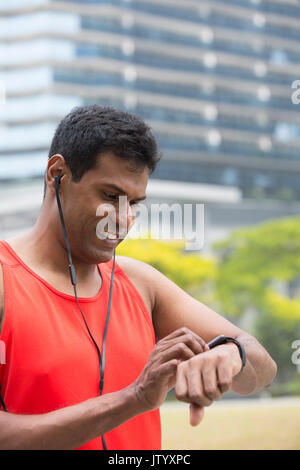 Sporty Indian man looking at his smartwatch heart rate monitor. Athletic Asian man using a smart watch and listening to music with earphones. Stock Photo