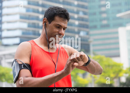 Sporty Indian man looking at his smartwatch heart rate monitor. Athletic Asian man using a smart watch and listening to music with earphones. Stock Photo