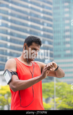 Sporty Indian man looking at his smartwatch heart rate monitor. Athletic Asian man using a smart watch and listening to music with earphones. Stock Photo