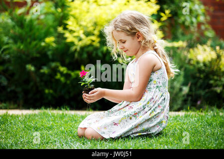 Little blonde girl holding young flower plant in hands on green background. Stock Photo