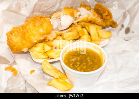 Traditional British takeaway meal of battered fish and chips, plus curry sauce in the containers and paper wrapping. England, UK. Stock Photo