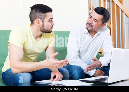 Two cheerful men sitting at the table and looking through laptop. Focus on the right man Stock Photo