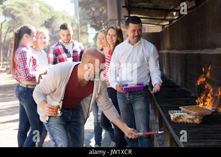 Cheerful adult group of friends relaxing at grill party at park. Focus on the man in the red t-shirt Stock Photo