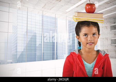 Girl balancing books and apple on head against modern room overlooking city Stock Photo