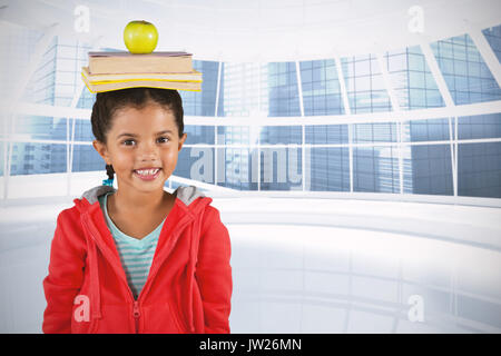 Smiling girl balancing books and apple on head against modern room overlooking city Stock Photo