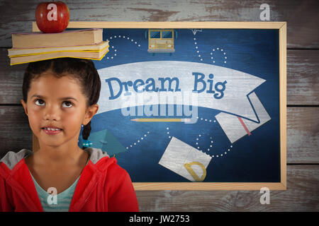 Girl balancing books and apple on head against dream big against blue chalkboard Stock Photo
