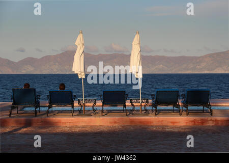 Silhouettes of two people sitting in beach chairs looking out over the deep blue Sea of Cortez and distant Isla Cerralvo in late golden evening sun. Stock Photo