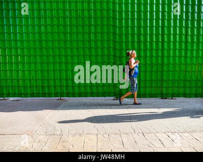 A middle-aged female Hispanic walks beside a bright green wall made of many tiles yielding a repetition of form in Havana. Stock Photo