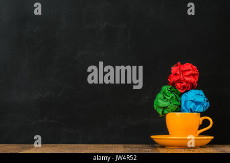 smooth glossy ceramic orange coffee cup put on the wooden surface table with color paper balls stacked together showing blackboard background. Stock Photo