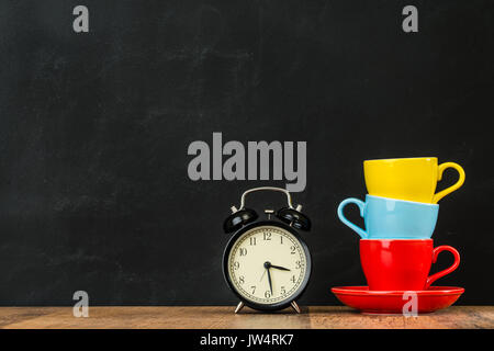 stacked colored coffee cups on vintage wood floor with alarm clock equipment showing design inspired copyspace on blackboard wall background in fashio Stock Photo
