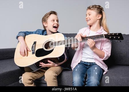 cheerful brother and sister playing on guitar and singing together while sitting on sofa Stock Photo