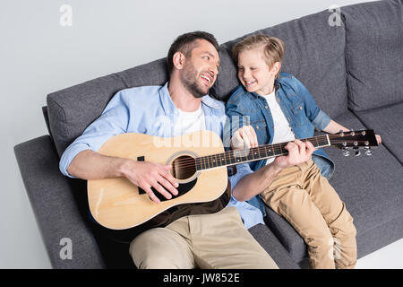 father playing on guitar and looking on his son while sitting on sofa Stock Photo